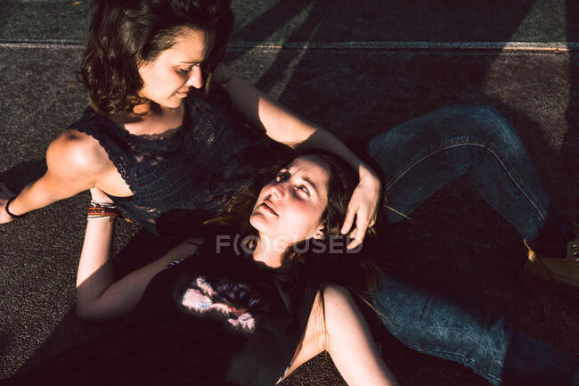 From above of dreamy female lovers in casual clothes sitting on asphalt ground looking at each other — Stock Photo