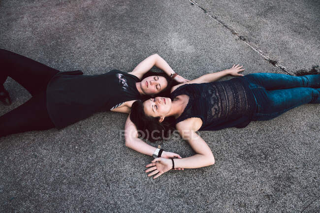 From above of dreamy female lovers in casual clothes lying on asphalt ground head to head with eyes closed — Stock Photo
