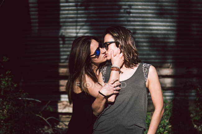 Tender female lovers wearing casual outfit and sunglasses kissing each other while walking outside on blurred street background in sunny summer day — Stock Photo