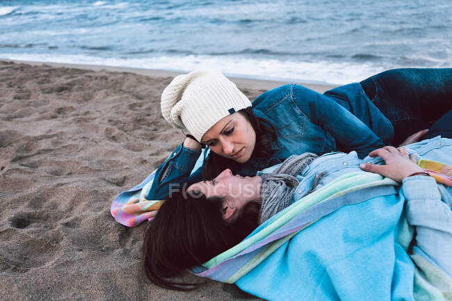 Conteúdo jovens amantes do sexo feminino deitados na praia de areia perto de água calma e olhando uns para os outros enquanto desfrutam de encontro passar tempo juntos — Fotografia de Stock
