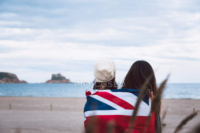 Visão traseira de amantes do sexo feminino irreconhecível envolvendo juntos na bandeira da Grã-Bretanha, enquanto abraçando contra a costa do mar e céu nublado durante a data romântica — Fotografia de Stock
