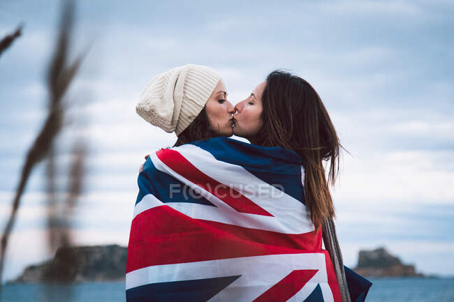 Vista lateral de amantes femeninos envueltos en la bandera de Gran Bretaña mientras se abrazan y besan con los ojos cerrados contra la costa del mar y el cielo nublado durante el fin de semana romántico - foto de stock