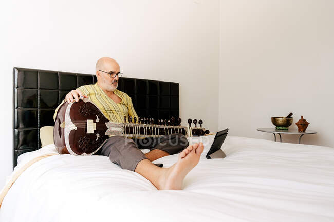 Elderly man sitting on bed with sitar and taking notes in notebook while watching online lesson on tablet — Stock Photo