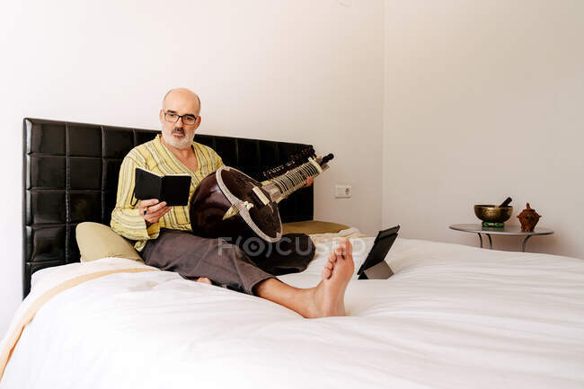 Älterer Mann sitzt mit Sitar im Bett und macht sich Notizen in Notizbuch, während er Online-Unterricht auf dem Tablet schaut — Stockfoto