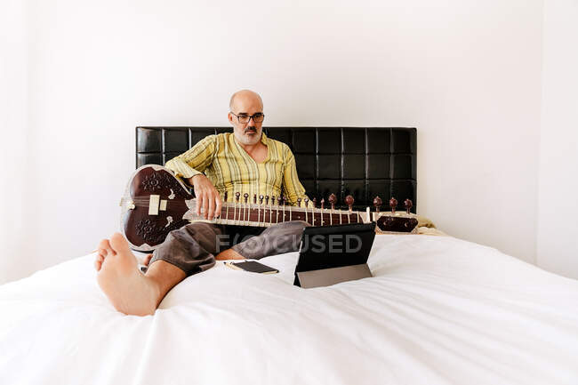 Літній чоловік сидить на ліжку з ситаром і записує нотатки в зошиті, спостерігаючи онлайн урок на планшеті. — стокове фото