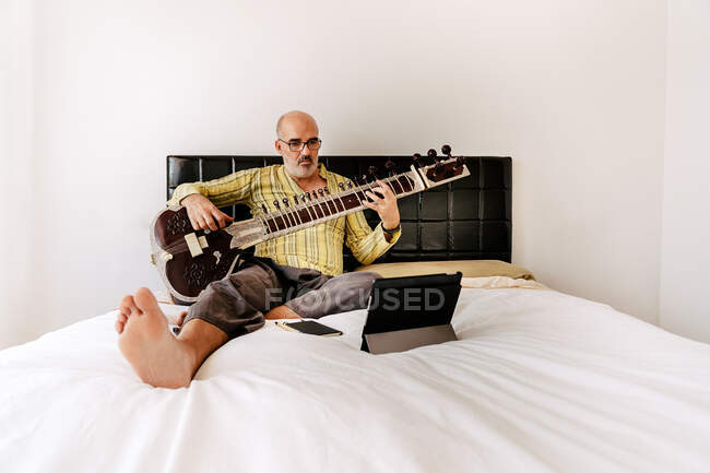 Elderly man sitting on bed with sitar and taking notes in notebook while watching online lesson on tablet — Stock Photo