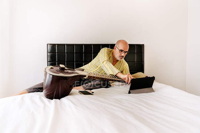 Homme âgé assis sur le lit avec sitar et de prendre des notes dans le carnet tout en regardant leçon en ligne sur tablette — Photo de stock