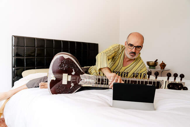 Uomo anziano seduto sul letto con sitar e prendere appunti nel notebook mentre si guarda lezione online su tablet — Foto stock
