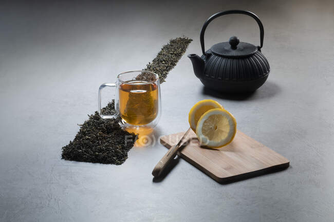 Aromatisches Getränk in Glasbecher und Teekanne mit Zitronen und Haufen getrockneter Teeblätter auf Tisch auf schwarzem Hintergrund — Stockfoto
