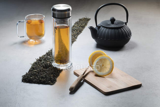 Ароматичний напій у скляному кухлі та чайнику, прикрашений лимонами та купою висушеного чайного листя на чорному тлі. — стокове фото