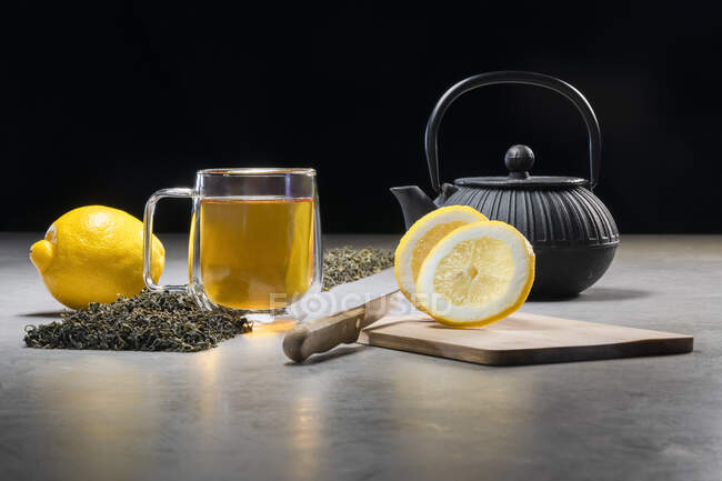 Boisson aromatique dans une tasse en verre et une théière disposées avec des citrons et des tas de feuilles de thé séchées sur la table sur fond noir — Photo de stock