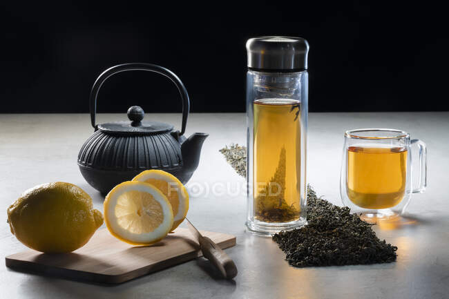 Aromatic beverage in glass mug and teapot arranged with lemons and heaps of dried tea leaves on table on black background — Stock Photo