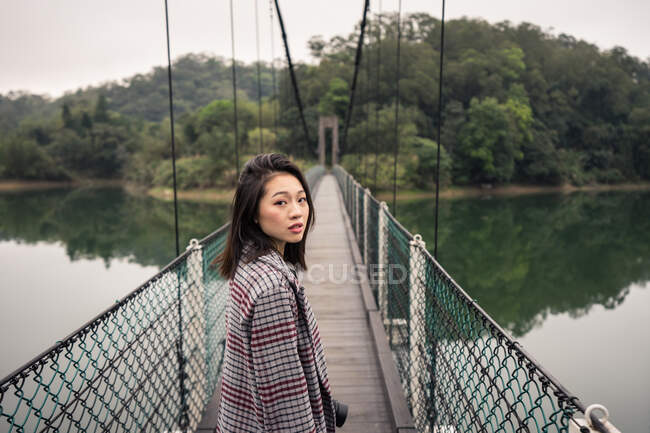 Vista lateral de turista asiática sonhadora em desgaste casual com câmera de foto em pé na ponte suspensa sobre o lago perto da vasta floresta e olhando para a câmera — Fotografia de Stock