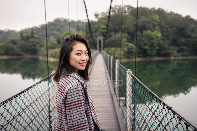 Vista lateral de la alegre turista asiática en ropa casual con cámara de fotos de pie en el puente colgante sobre el lago cerca de un vasto bosque y mirando a la cámara - foto de stock