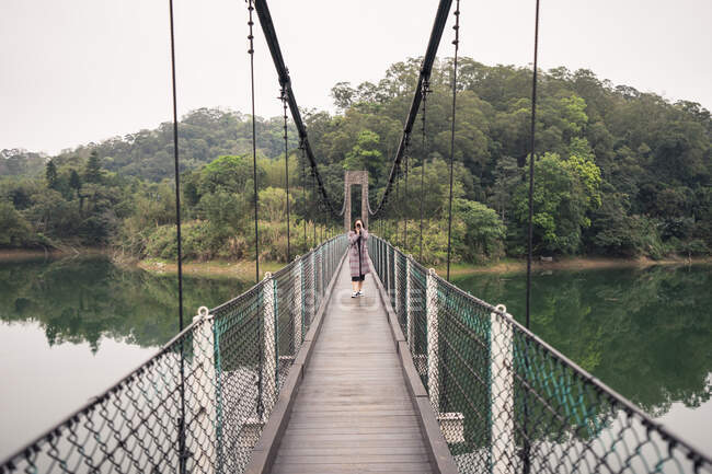 Voyageuse en manteau prenant des photos avec appareil photo debout sur un pont suspendu en bois avec clôture métallique au-dessus d'un lac près d'une forêt verte — Photo de stock