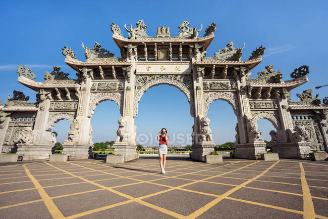 Снизу тонкая женщина-путешественница фотографируется с фотоаппаратом, стоя на тротуаре возле фасада китайского храма с арками, украшенными скульптурами — стоковое фото