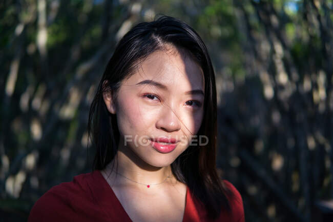 Pensativo joven mujer asiática en ropa casual y pequeño collar mirando a la cámara mientras está de pie en el bosque a la luz del día - foto de stock