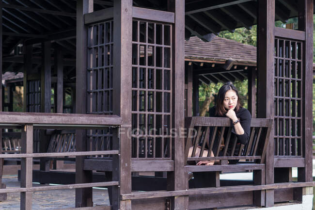 Мрія молода азіатка сидить на дерев'яній лавці на огородженій терасі біля дерев, спираючись на руку і дивлячись у денне світло — стокове фото