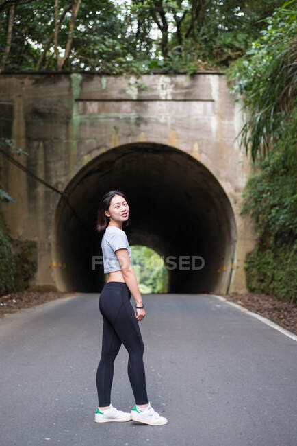 Vue latérale d'une jeune femme asiatique mince en legging debout sur une route asphaltée en face d'un tunnel près d'un mur recouvert de plantes vertes et regardant la caméra — Photo de stock