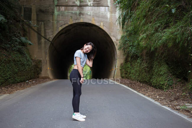 Vista lateral de delgada hembra asiática joven en leggings de pie en la carretera de asfalto frente al túnel cerca de la pared cubierta de plantas verdes y mirando a la cámara - foto de stock
