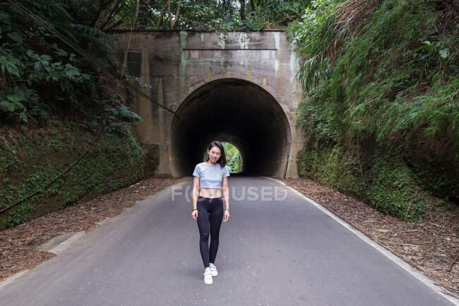 Вид сбоку на стройную молодую азиатку в леггинсах на асфальтированной дороге перед туннелем возле стены, покрытой зелеными растениями и идущую к камере — стоковое фото
