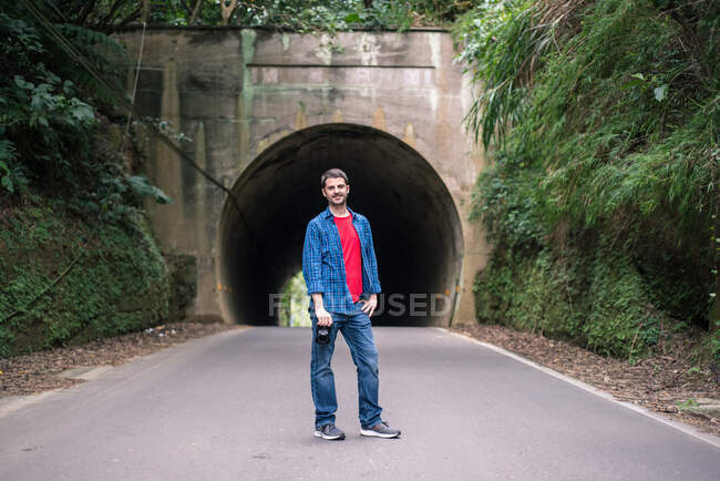 Чоловічий турист у червоній сорочці та джинсовій куртці стоїть з цифровою фотокамерою на асфальтній дорозі поблизу зеленого кріплення та тунелю в денний час — стокове фото