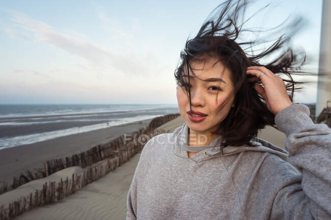 Calme jeune Asiatique femelle en tenue décontractée toucher les cheveux et regarder la caméra tout en se tenant près de la côte sous le ciel nuageux par temps venteux — Photo de stock