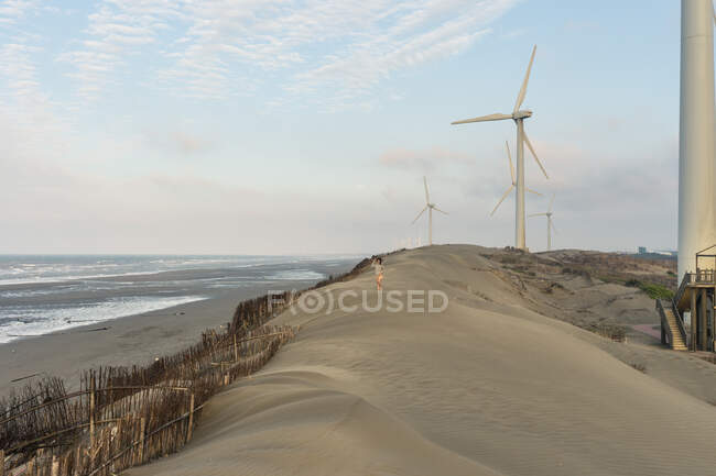 Anonymous person walking on sand hill near sea beach and windmills with high tower under cloudy sky in afternoon — Stock Photo