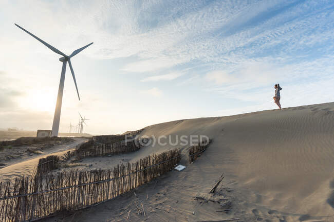 De baixo vista lateral de turista masculino anônimo em pé na encosta de areia perto moinhos de vento e cerca feita de galhos de árvore sob céu nublado — Fotografia de Stock