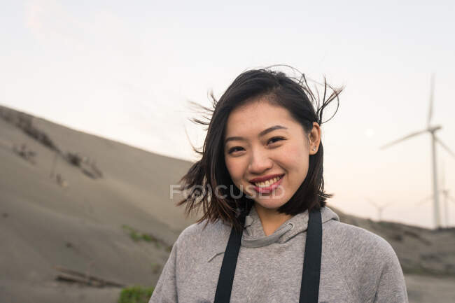 Calma giovane asiatica femmina in abbigliamento casual toccare i capelli e guardando la fotocamera mentre in piedi vicino al mare sotto il cielo nuvoloso il giorno ventoso — Foto stock
