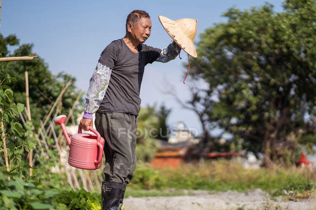 Seitenansicht eines asiatischen Mannes in schmutziger Kleidung mit orientalischem Strohhut, der Plastik-Gießkannen zum Ausgießen von Pflanzen füllt, während er neben einem schmalen Bach auf einer Farm in Taiwan steht — Stockfoto
