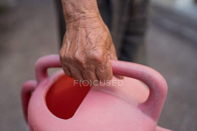 De arriba persona étnica irreconocible con pote de riego rojo malhumorado de pie sobre fondo gris borroso mientras trabajaba en el jardín en Taiwán - foto de stock