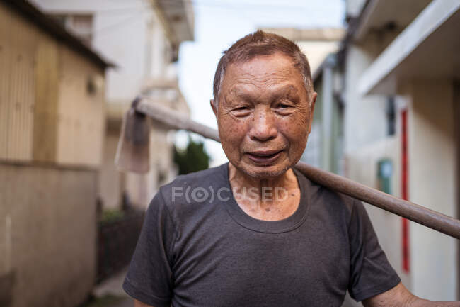 Portrait of elderly Asian male gardener in casual clothes smiling at camera while standing on street with hoe over shoulder in settlement in Taiwan — Stock Photo
