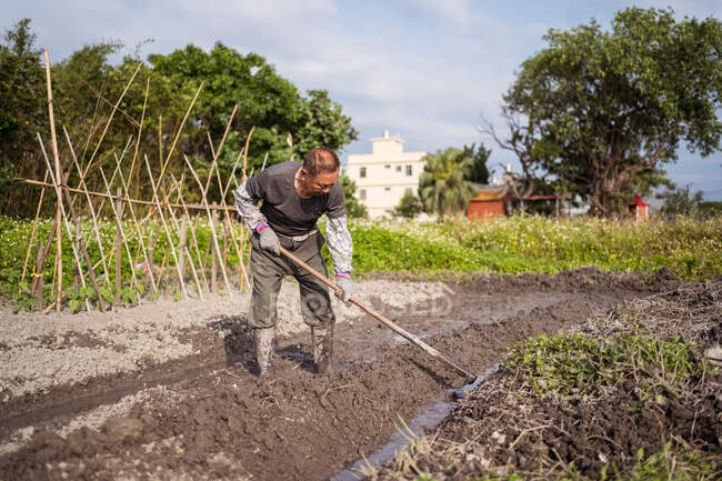 Focado homem asiático em roupas casuais e botas cultivando solo molhado usando enxada antes de plantar no jardim em Taiwan — Fotografia de Stock