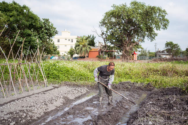 Focado homem asiático em roupas casuais e botas cultivando solo molhado usando enxada antes de plantar no jardim em Taiwan — Fotografia de Stock