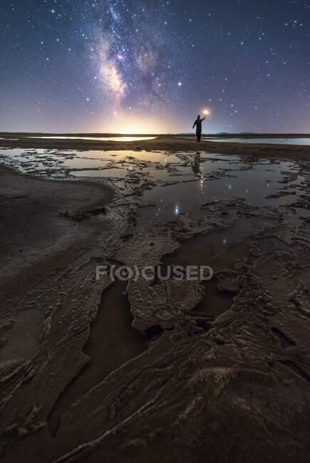 Back view of anonymous man standing and holding torch on empty road among calm water and reaching out to star under colorful nigh sky with milky way on background — Stock Photo