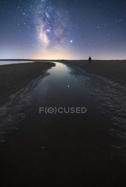 Vista posterior del hombre anónimo de pie en el camino vacío entre el agua tranquila y llegar a la estrella bajo el colorido cielo cercano con la vía láctea en el fondo - foto de stock
