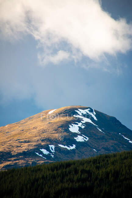 Вид на гору, вкритий снігом серед зелених пагорбів з лісом проти хмарного неба навесні в Шотландському нагір 