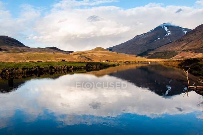 Increíble paisaje escocés de lago tranquilo con superficie espejada que refleja la montaña con pico cubierto de nieve y cielo azul nublado en la zona de Glen Coe - foto de stock