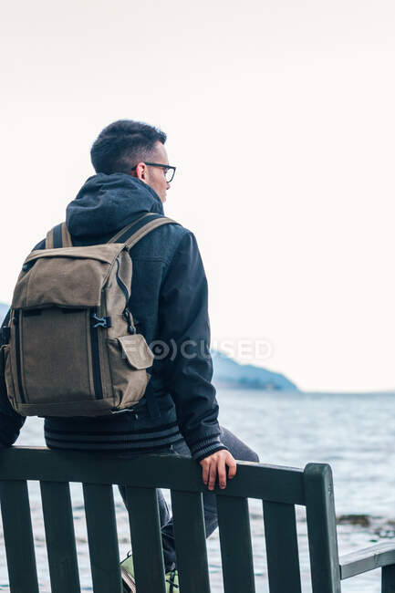 Rückenansicht eines männlichen Wanderers in warmer Jacke mit Rucksack, der auf einer Holzbank am Meer sitzt und die Meereslandschaft mit felsiger Küste während einer Reise in Schottland genießt — Stockfoto