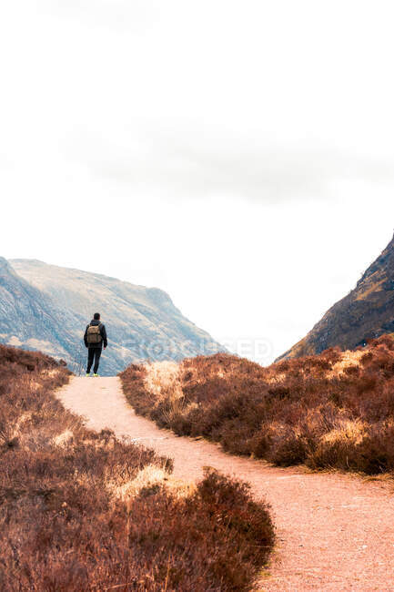Vista trasera de excursionista irreconocible con mochila de pie en el sendero que conduce a través de colinas con hierba seca y admirando la vista de las montañas contra el cielo nublado en las tierras altas escocesas - foto de stock