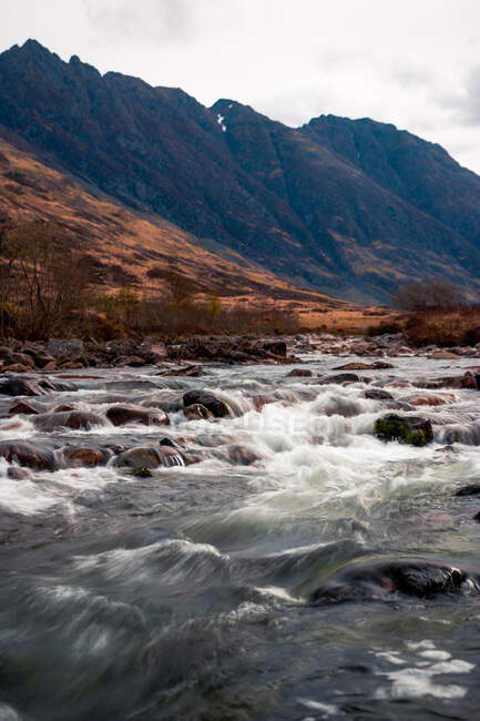 Paisaje natural áspero con río de montaña que fluye entre las tierras altas rocosas en un día nublado en Escocia - foto de stock