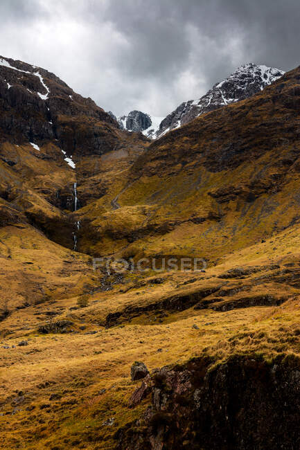 Picturesque view of mountain slope covered with snow among green hills with forest against cloudy sky in spring day in Scottish Highlands — Stock Photo