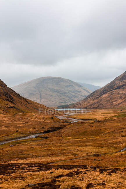 Estrada curvilínea estreita que atravessa terreno montanhoso com grama seca entre montanhas rochosas no dia nublado da primavera nas Terras Altas da Escócia — Fotografia de Stock