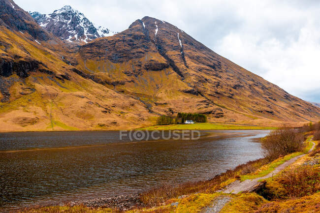 Increíble paisaje escocés de lago tranquilo con superficie espejada que refleja la montaña con pico cubierto de nieve y cielo azul nublado en la zona de Glen Coe - foto de stock