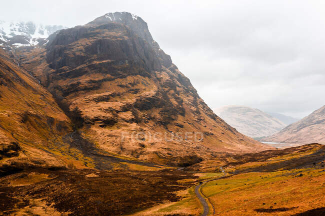 Узкая извилистая дорога проходит по холмистой местности с сухой травой среди скалистых гор в облачный весенний день в Шотландском нагорье — стоковое фото
