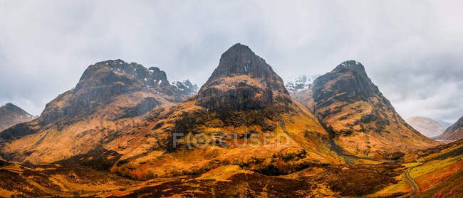 Stretta strada sinuosa che attraversa terreni collinari con erba secca tra le montagne rocciose nella nuvolosa giornata primaverile nelle Highlands scozzesi — Foto stock