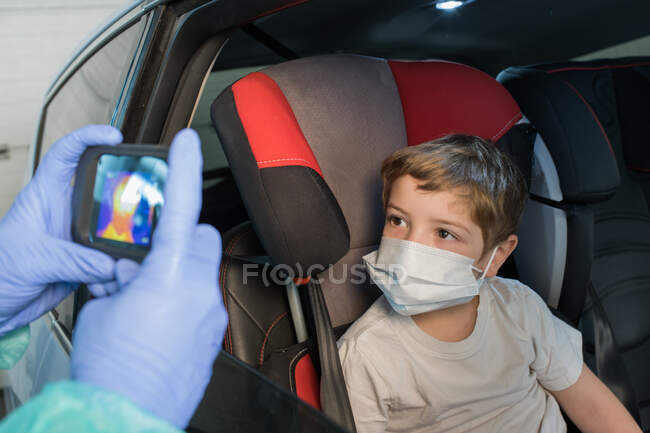 Médico de la cosecha en guantes médicos que miden la temperatura del niño mientras usa la cámara infrarroja durante la epidemia de coronavirus - foto de stock