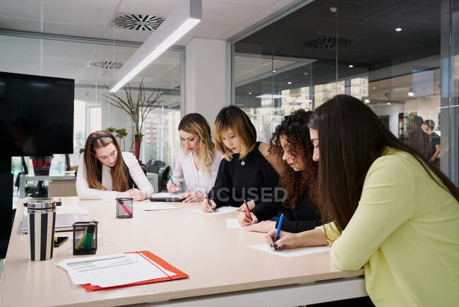 Группа молодых многорасовых коллег-женщин в повседневной одежде собирается вокруг ноутбука и наблюдает за деловой информацией, работая вместе в современном рабочем пространстве — стоковое фото