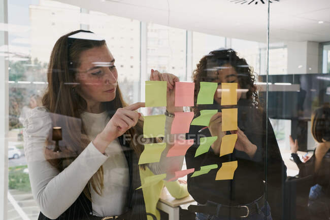 Серйозна молода багатонаціональна жінка-колеги стоять поруч зі скляною стіною з липкими нотатками і аналізують деталі нового проекту, одночасно обмінюючись ідеями і мозком в сучасному робочому місці. — стокове фото